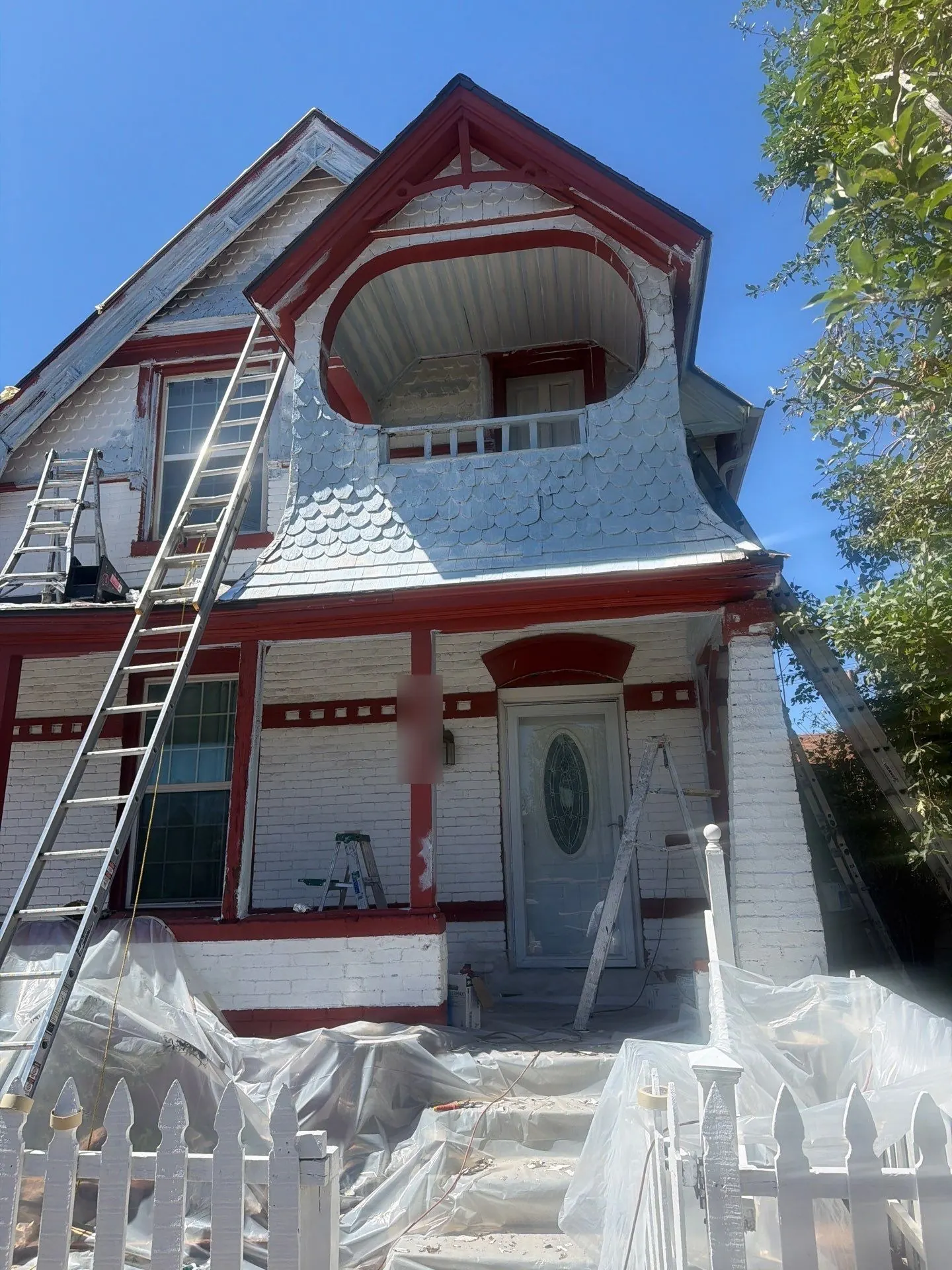 Plastic sheeting laid over the porch steps and ground of a historic Denver Victorian during lead-safe exterior paint prep, with scraped fish-scale shingles above